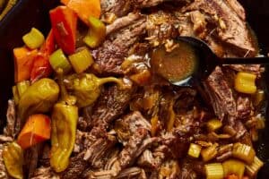 Close-up of shredded Mississippi pot roast in a dark pot with cooked carrots, celery, pepperoncinis, and gravy