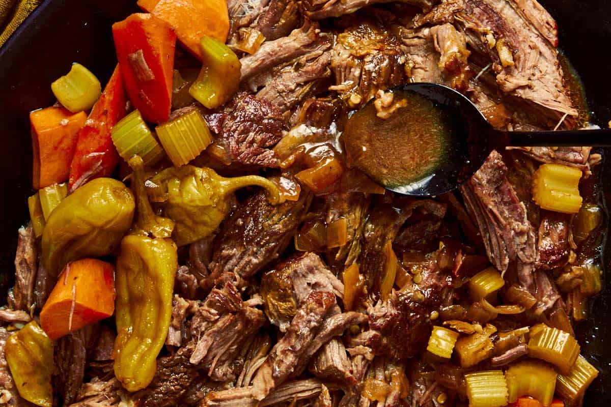 Close-up of shredded Mississippi pot roast in a dark pot with cooked carrots, celery, pepperoncinis, and gravy