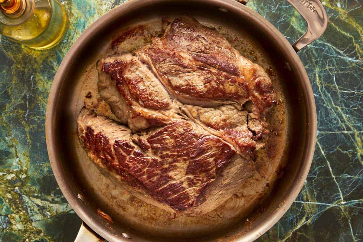 A beef chuck roast fully seared and browned in a stainless steel skillet over a green marble surface.