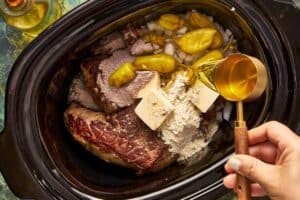 A top-down view of a slow cooker containing a seared beef roast, diced onions, pepperoncini peppers, butter pats, and two types of dry seasoning powder, while a hand pours liquid from a metal measuring cup over the ingredients.