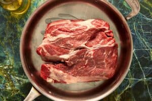 A large, raw beef chuck roast sitting in a stainless steel skillet with cooking oil, ready to be seared.