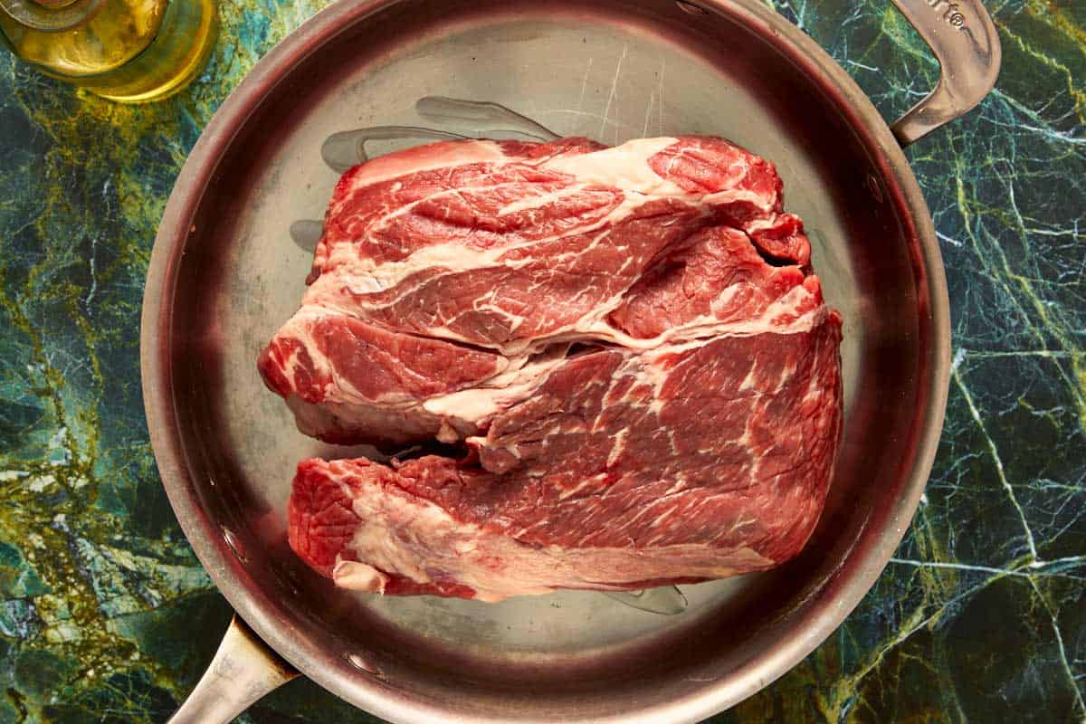 A large, raw beef chuck roast sitting in a stainless steel skillet with cooking oil, ready to be seared.