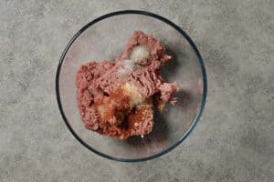 A glass bowl containing raw ground turkey topped with dry spices ready to be mixed for a turkey burger recipe.