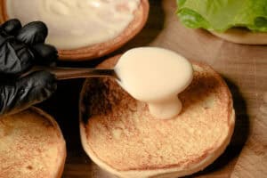 A hand wearing a black glove uses a metal spoon to spread creamy hot honey aioli onto a toasted hamburger bun.