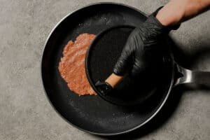A hand wearing a black glove uses a heavy wooden-handled burger press to flatten a raw turkey meatball in a hot black skillet, demonstrating the smash burger technique.