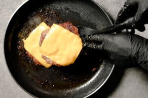 Hands wearing black gloves use a spatula to stack two cooked patties topped with melted cheese in a hot skillet while making turkey smash burgers.
