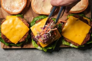 Tongs placing coleslaw onto a burger patty topped with melted cheese and lettuce, with sauced buns in the background.