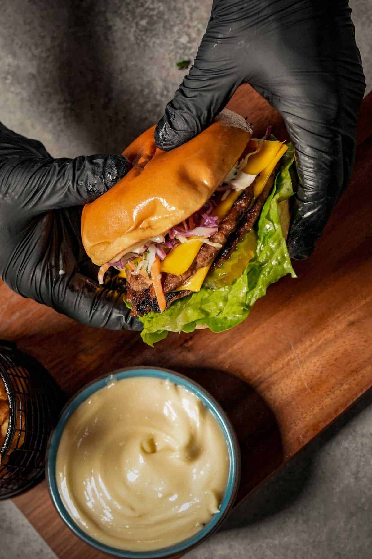 Hands wearing black gloves holding an assembled turkey burger with a bowl of hot honey aioli in the background, showcasing the final result a smash burger recipe.