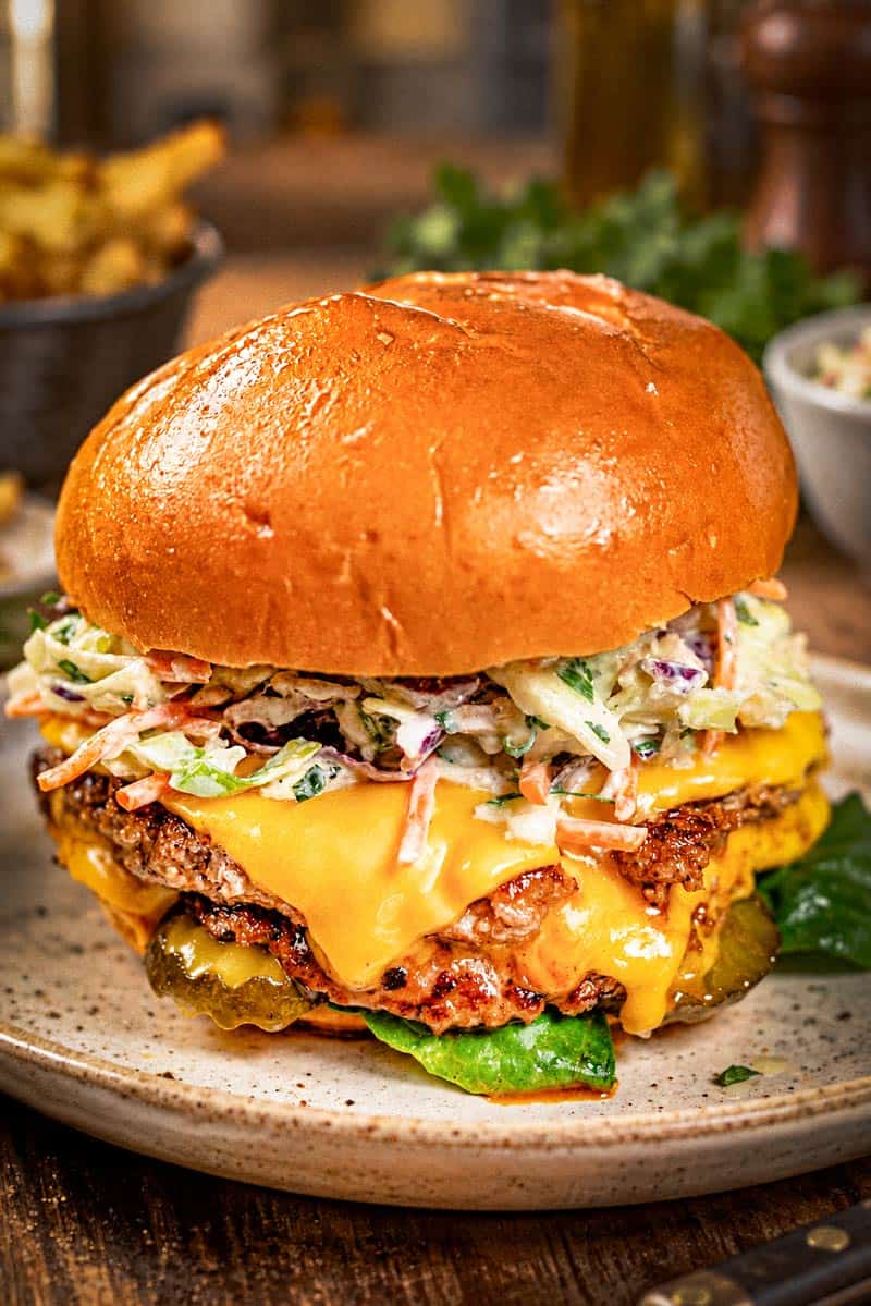 A close-up of a stacked turkey smash burger on a toasted brioche bun, featuring double patties with melted cheddar cheese, creamy coleslaw, sliced pickles, and lettuce.
