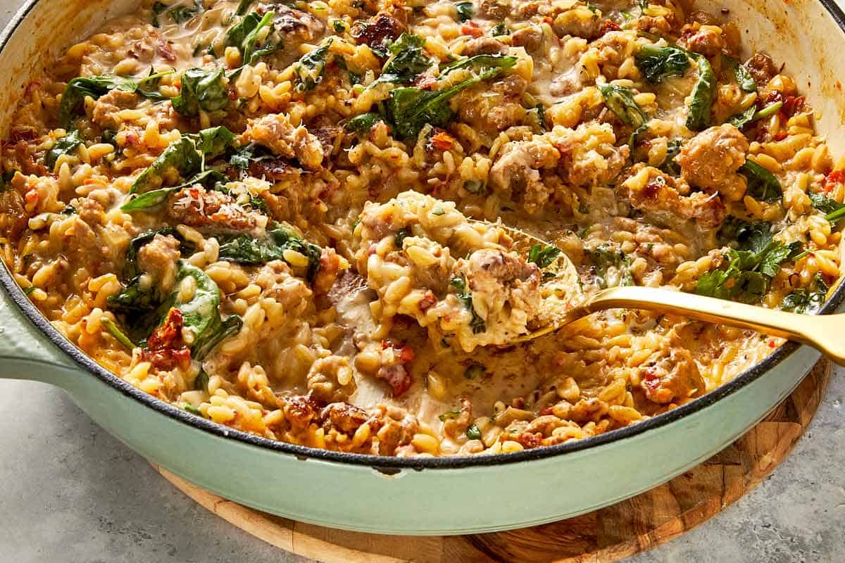 Close-up of a gold spoon scooping creamy Italian sausage and spinach orzo pasta from a light green enameled cast-iron skillet.   