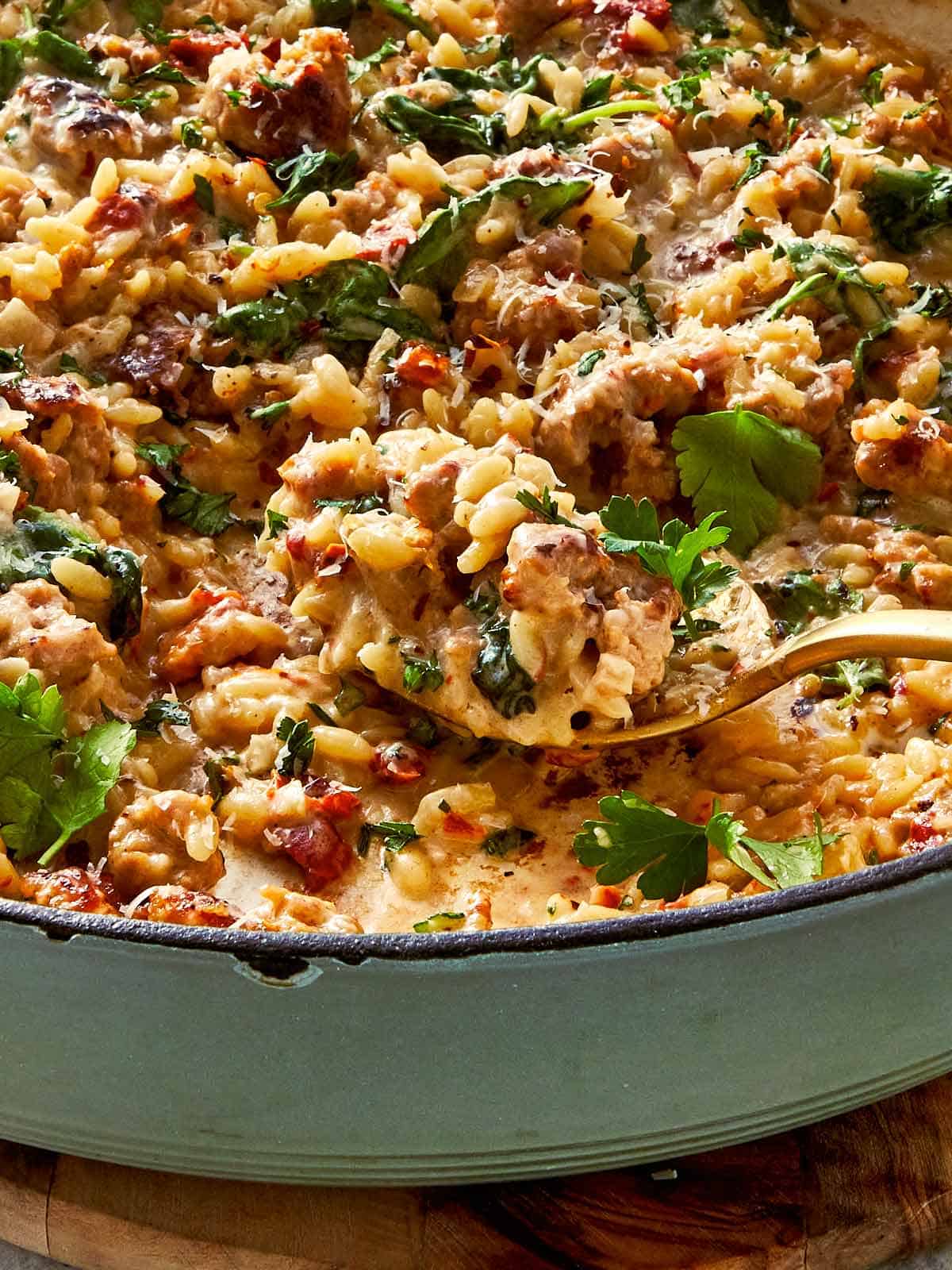 Gold spoon scooping creamy Italian sausage and spinach orzo pasta from a light green enameled cast-iron skillet.