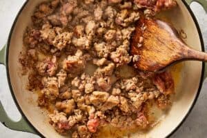 Overhead view of Italian sausage being broken up and browned in an enameled cast-iron skillet with a wooden spatula.