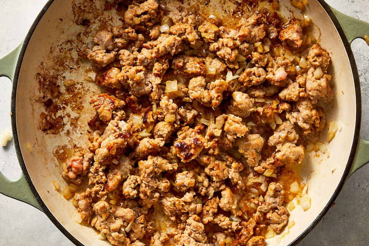 Overhead view of crumbled Italian sausage and diced onions browned in an enameled cast-iron skillet.