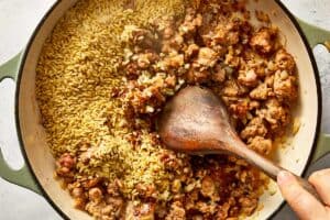 Overhead view of uncooked orzo pasta, minced garlic, and crushed red pepper flakes being stirred into a skillet of browned Italian sausage and onions with a wooden spoon.