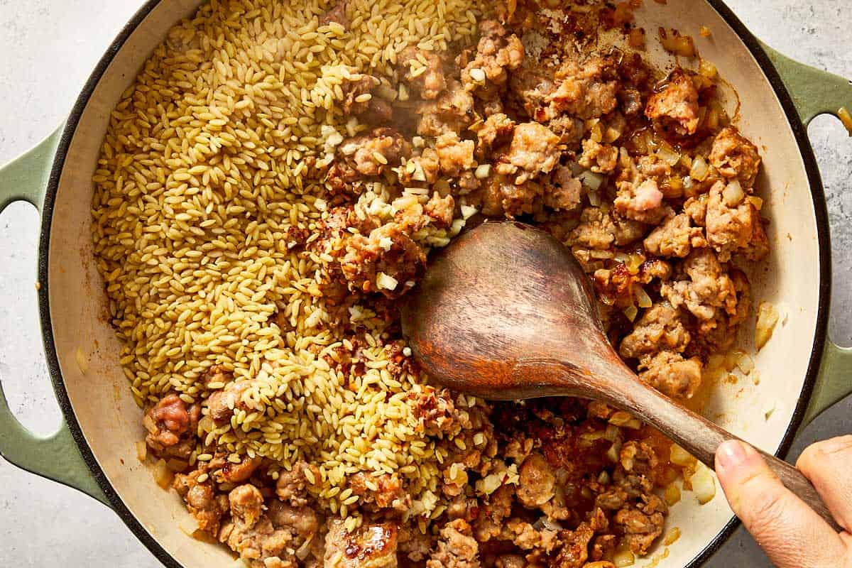 Overhead view of uncooked orzo pasta, minced garlic, and crushed red pepper flakes being stirred into a skillet of browned Italian sausage and onions with a wooden spoon.   