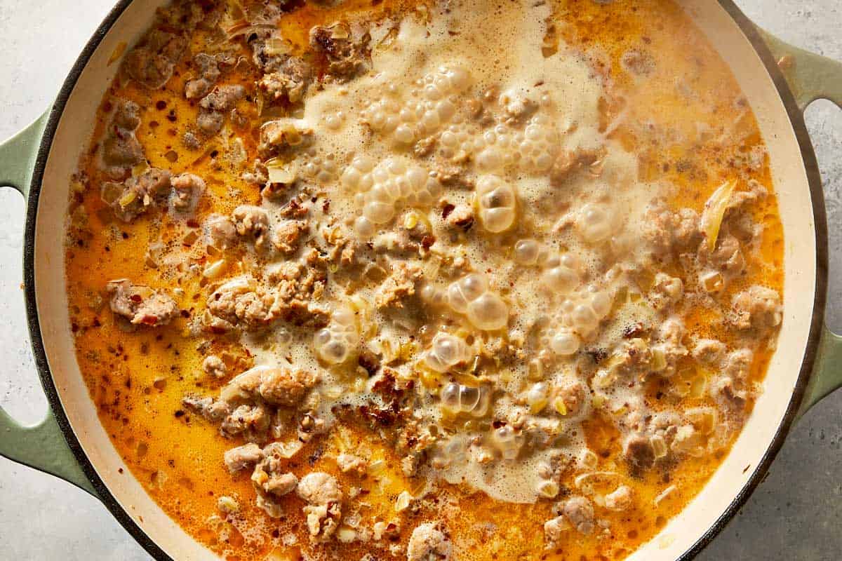 Overhead view of browned Italian sausage, diced onions, and orzo pasta simmering in chicken broth and cream in an enameled cast-iron skillet.