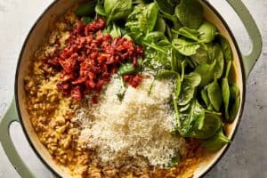 Overhead view of fresh baby spinach, chopped sun-dried tomatoes, and freshly grated Parmesan cheese added on top of cooked sausage and orzo in a skillet.