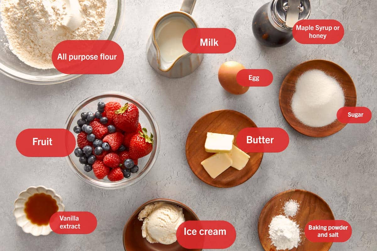 Overhead view of ingredients arranged on a grey surface, including bowls of all purpose flour, mixed berries, sugar, butter, vanilla extract, ice cream, baking powder, and salt, along with a pitcher of milk, a brown egg, and a jar of maple syrup, each labeled with a red tag.