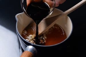 A hand pouring dark soy sauce from a small glass bowl into a saucepan containing honey and minced garlic, with a wooden spoon resting inside.