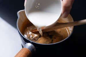 A hand pouring a white cornstarch slurry from a small white bowl into a simmering brown sauce in a saucepan.