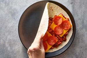 Overhead view of a hand folding the empty half of a flour tortilla over the pizza filling, cheddar cheese slices, and pepperoni slices.
