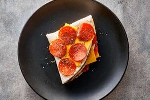 Overhead view of a folded triangular pizza tortilla wrap on a black plate, topped with a slice of cheddar cheese, five pepperoni slices, and dried Italian herbs before cooking.