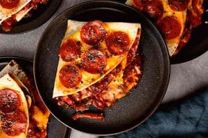 Overhead close-up of a folded triangular pepperoni pizza tortilla wrap on a black plate, topped with melted cheddar cheese, crispy pepperoni slices, and dried herbs, with rich red tomato and mushroom filling oozing from the side.