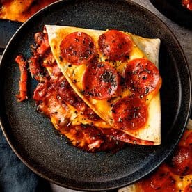 Overhead close-up of a folded triangular pepperoni pizza tortilla wrap on a black plate, topped with melted cheddar cheese, crispy pepperoni slices, and dried herbs, with rich red tomato and mushroom filling oozing from the side.