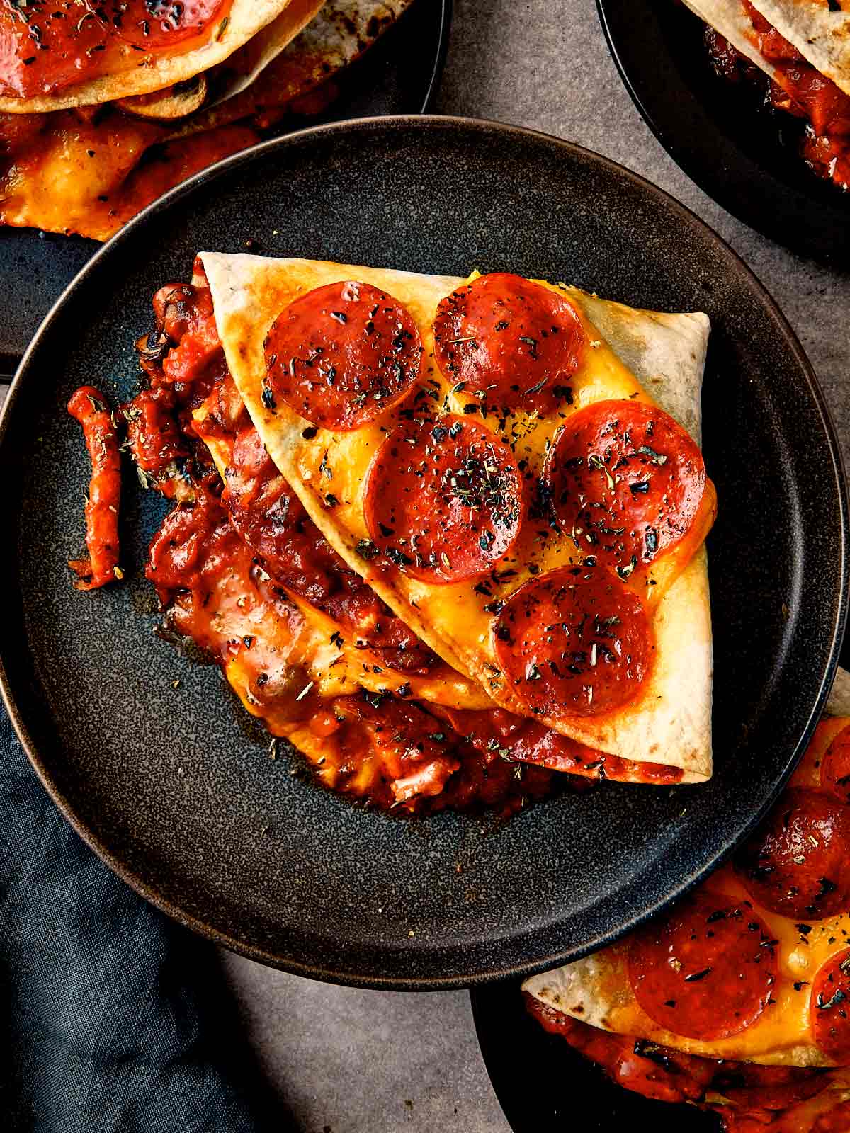 Overhead close-up of a folded triangular pepperoni pizza tortilla wrap on a black plate, topped with melted cheddar cheese, crispy pepperoni slices, and dried herbs, with rich red tomato and mushroom filling oozing from the side.