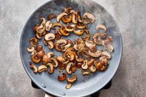 Overhead view of sautéed brown mushrooms in a skillet topped with shredded ham, minced garlic, dried Italian herbs, and red spices.