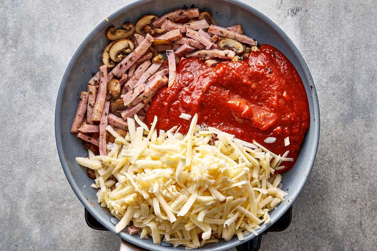 Overhead view of a skillet containing sautéed mushrooms, shredded ham, red pizza sauce, and unmelted shredded cheese being stirred together with a silicone spoon.