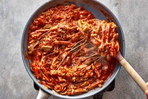 Overhead view of cooked pizza wrap filling in a skillet, with a spoon lifting a scoop of the hot, cheesy mixture of ham, mushrooms, and tomato sauce to show long strands of melted cheese.
