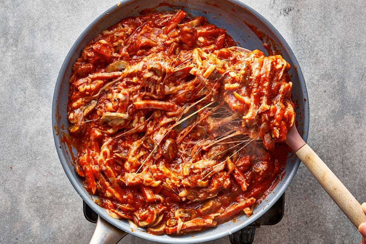 Overhead view of cooked pizza wrap filling in a skillet, with a spoon lifting a scoop of the hot, cheesy mixture of ham, mushrooms, and tomato sauce to show long strands of melted cheese.