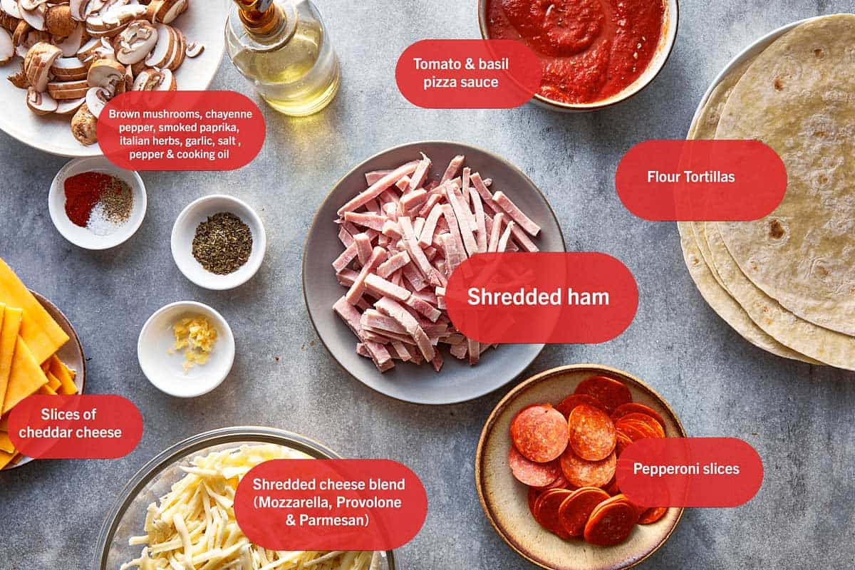 Overhead flat-lay photo of labeled pizza tortilla wrap ingredients in bowls and plates on a gray countertop: sliced brown mushrooms, spices, cooking oil, tomato and basil pizza sauce, flour tortillas, shredded ham, pepperoni slices, cheddar cheese slices, and a shredded mozzarella, provolone, and parmesan cheese blend.