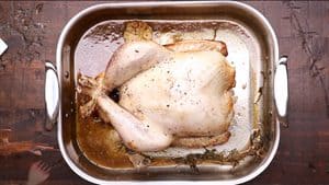 A top-down view of a partially cooked turkey with trussed legs resting in a stainless steel roasting pan, surrounded by pan juices, half a head of garlic, and sprigs of herbs on a dark wooden surface.