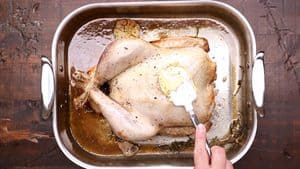 A top-down view of a hand using a white spatula to spread a dollop of butter over the breast of a partially cooked turkey in a stainless steel roasting pan, surrounded by pan juices, garlic, and herbs on a rustic wooden surface.