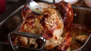 A close-up view of a beautifully browned, roasted turkey in a stainless steel pan being basted with its own savory pan juices using a large metal spoon.