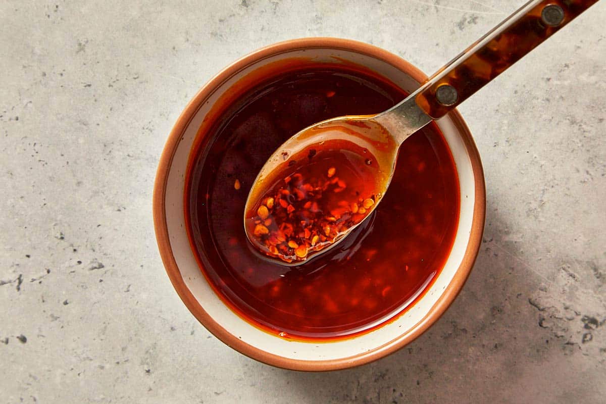 A small bowl of dark red dumpling sauce with a spoon resting inside, lifting some of the chili flakes from the mixture.