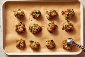 Raw shrimp dumpling filling scooped into small balls on a parchment-lined baking sheet, with a metal cookie scoop used for portioning.