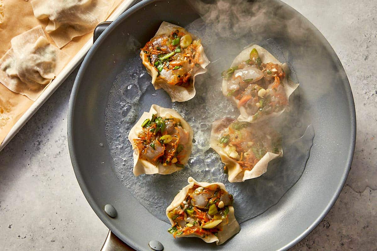 Assembled shrimp dumplings cooking in a hot skillet with bubbling water and steam, placed with the prawn filling facing up.