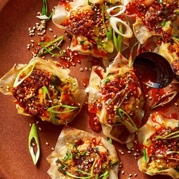 Top-down view of pan-fried shrimp dumplings on a brown plate, topped with chili oil sauce, sesame seeds, and sliced green onions, with a black spoon pouring more sauce over a dumpling on the right side.