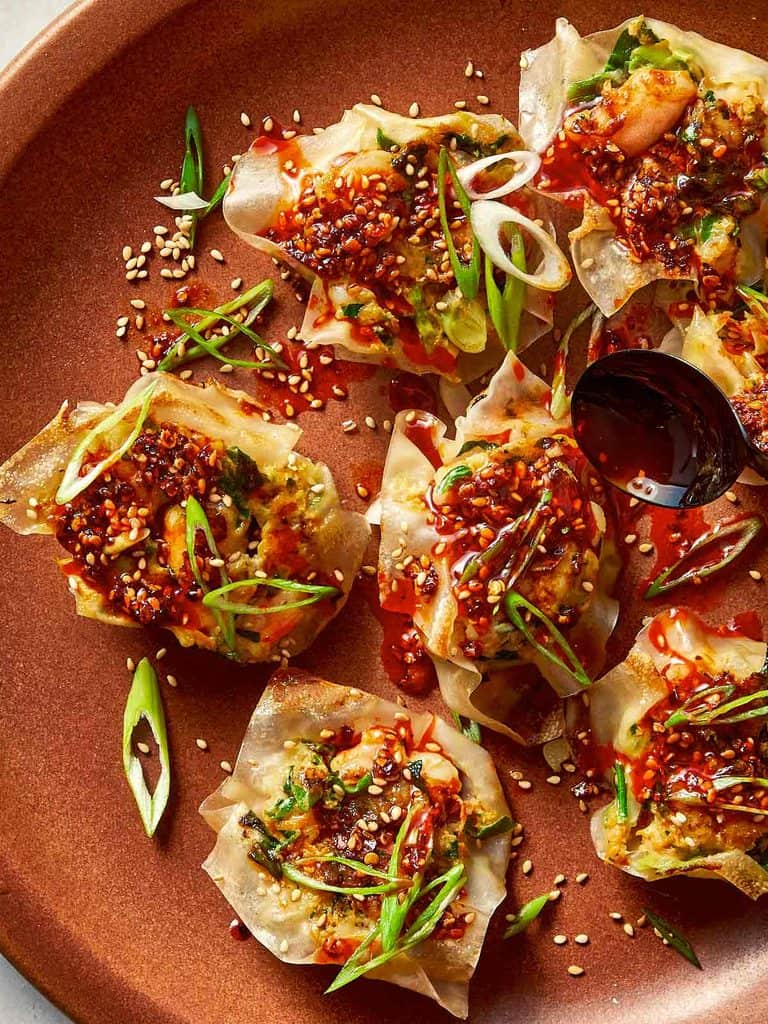 Top-down view of pan-fried shrimp dumplings on a brown plate, topped with chili oil sauce, sesame seeds, and sliced green onions, with a black spoon pouring more sauce over a dumpling on the right side.