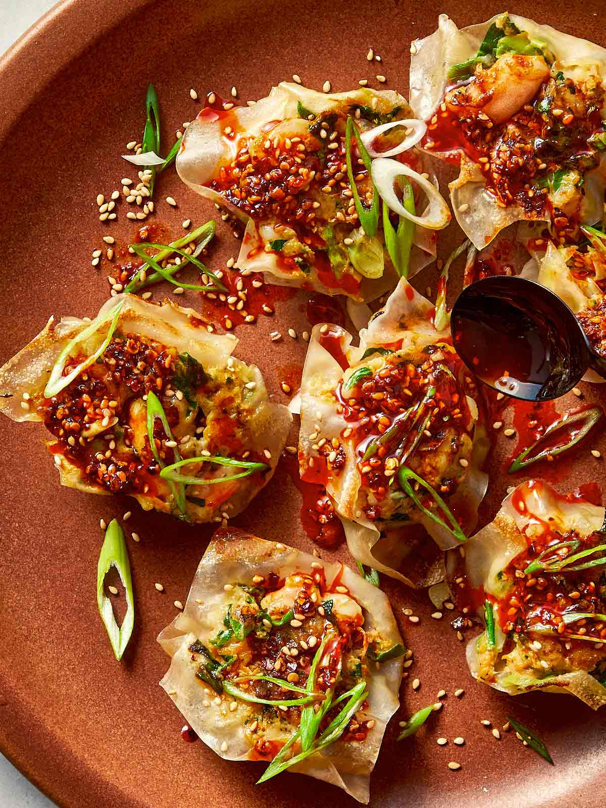 Top-down view of pan-fried shrimp dumplings on a brown plate, topped with chili oil sauce, sesame seeds, and sliced green onions, with a black spoon pouring more sauce over a dumpling on the right side.