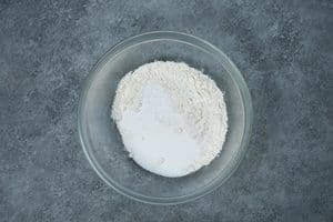 Waffles-1.jpg: A clear glass bowl on a textured grey surface containing unmixed white flour and sugar.