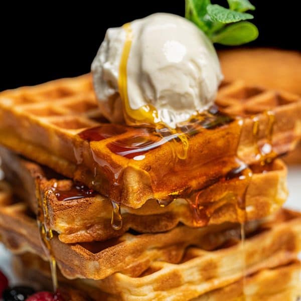 Stack of three waffles on a white plate, topped with a scoop of ice cream, a sprig of mint, and syrup dripping down the sides, accompanied by berries at the base.