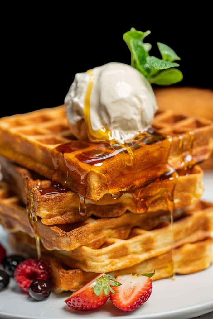 Stack of three waffles on a white plate, topped with a scoop of ice cream, a sprig of mint, and syrup dripping down the sides, accompanied by berries at the base.