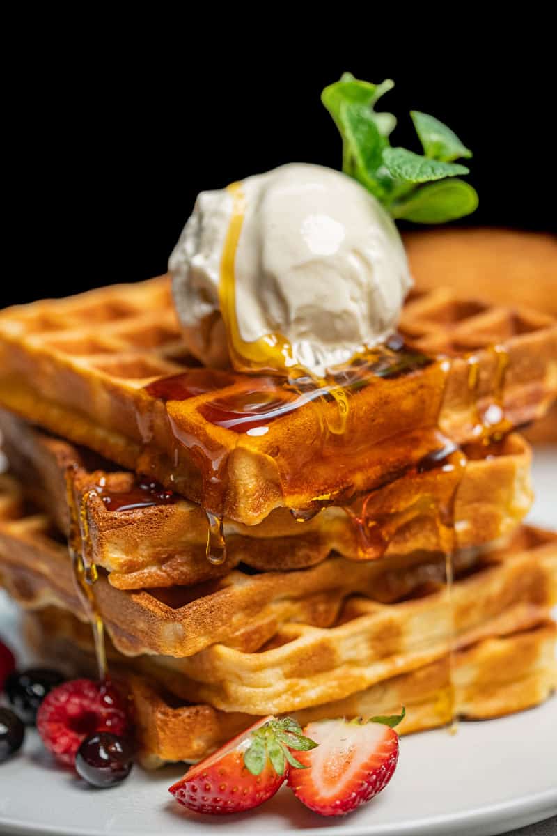 Stack of three waffles on a white plate, topped with a scoop of ice cream, a sprig of mint, and syrup dripping down the sides, accompanied by berries at the base.   