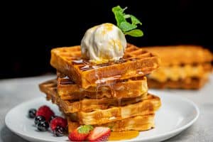 A stack of four square golden waffles on a white plate accompanied by fresh strawberries, raspberries, and blueberries.