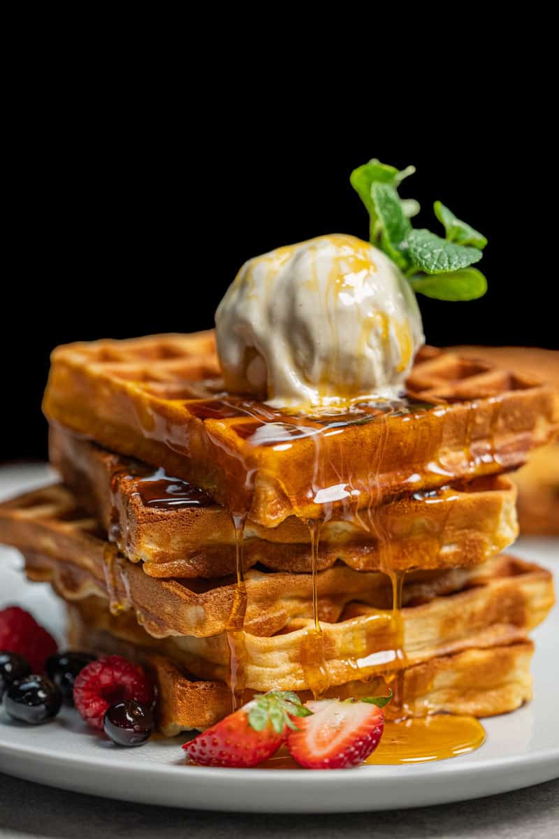 Stack of three waffles on a white plate, topped with a scoop of ice cream, a sprig of mint, and syrup dripping down the sides, accompanied by berries at the base.