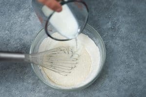 Milk being poured from a measuring cup into a glass bowl of dry ingredients with a metal whisk resting inside.